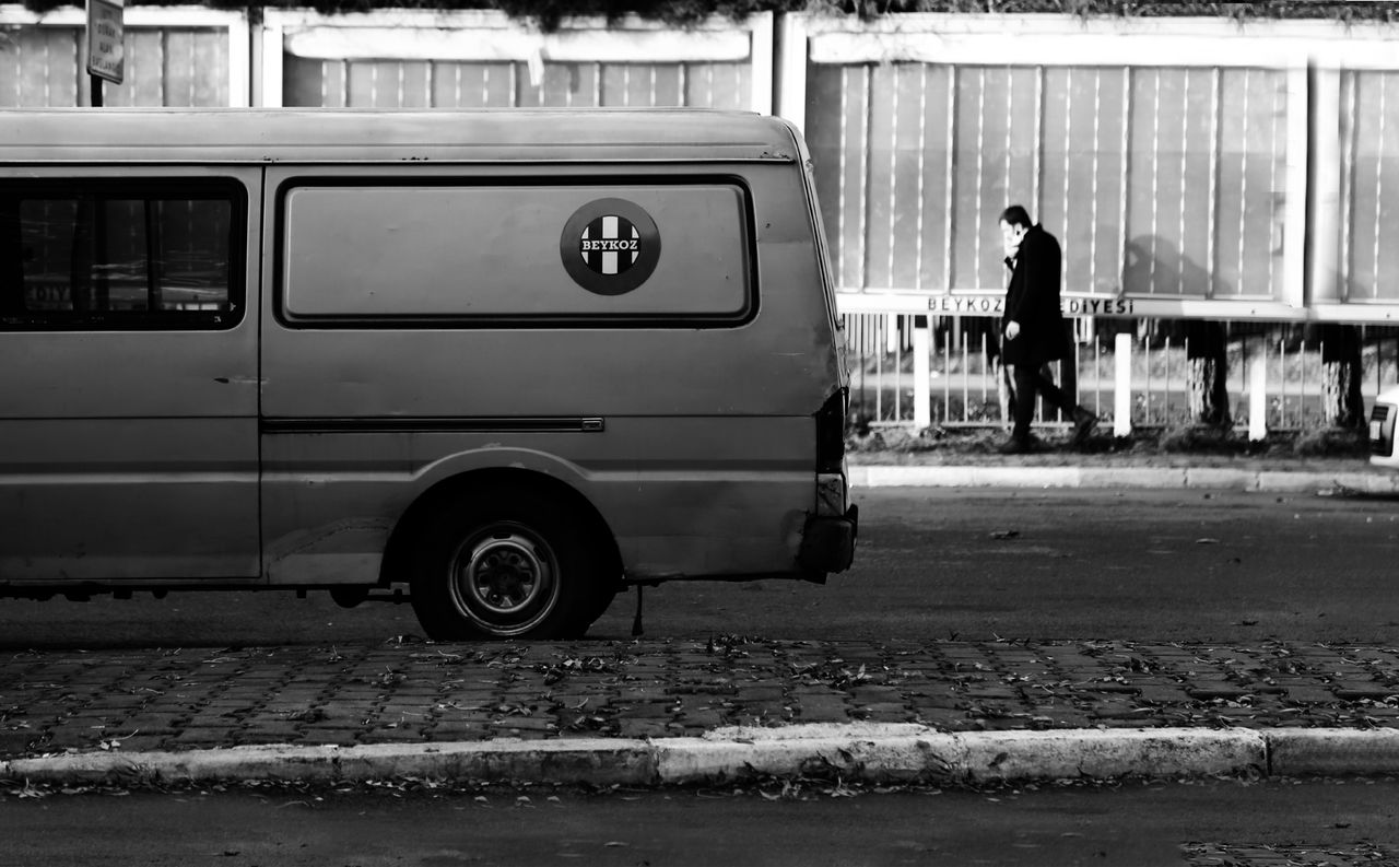 Black and white image of a parked van on a street with a person walking by, conveying urban life.