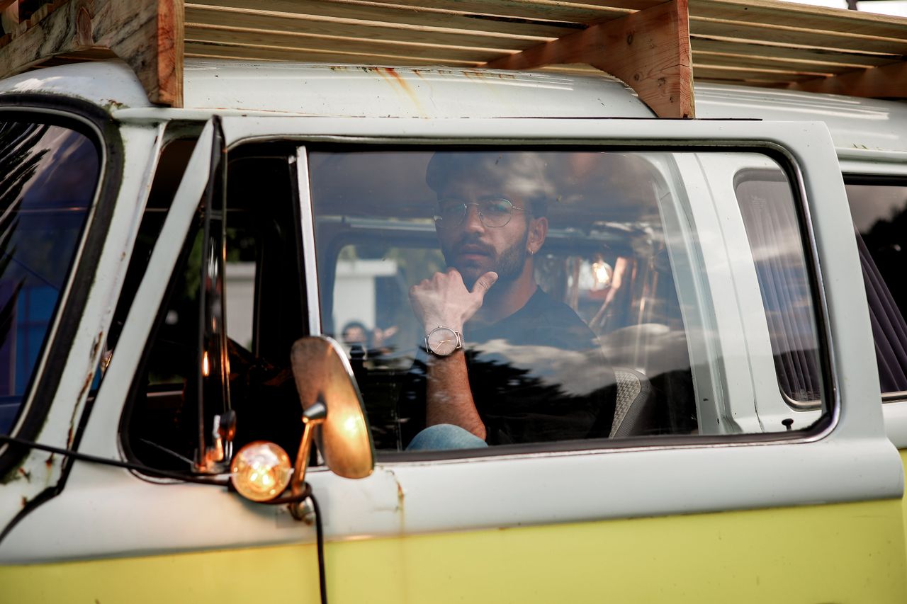 A man wearing eyeglasses sitting inside a classic vintage van, looking contemplative.