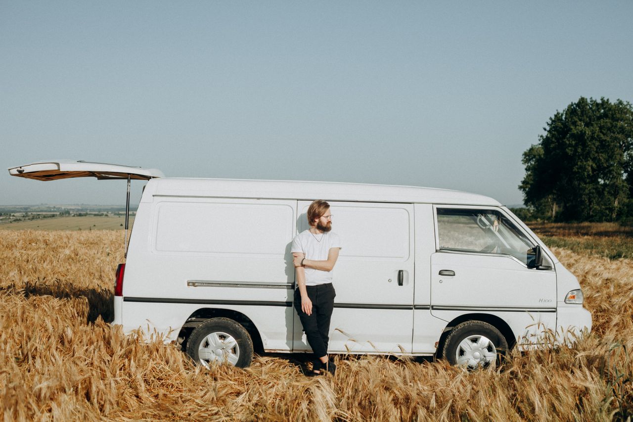 Bearded man in casual wear leaning on a van in a sunny rural landscape.
