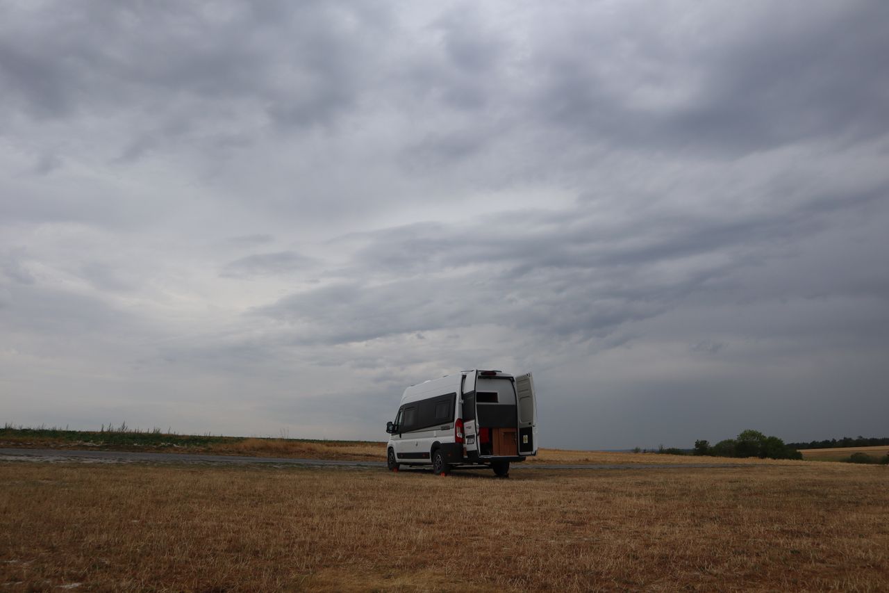 Camper van parked in open farmland under a dramatic, cloudy sky in Massiges, France.